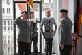 Oberst Timm steht im Festsaal und salutiert vor Generalmajor Henne. Im Hintergrund vor dem Fenster sind zwei Soldaten mit der Fahne des Landskommandos zu sehen.