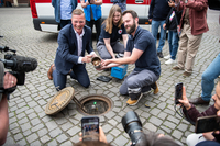 Frank Imhoff und ein Mitarbeiter des Roten Kreuzes knien vor dem geöffneten Bremer Loch. Zwischen sich halten die einen mit Münzen gefüllten Metallzylinder.