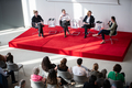 Vier Frauen sitzen auf einem Podium im Festsaal, vor ihnen sind mit Gästen gefüllte Stuhlreihen zu sehen.