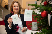 Antje Grotheer behängt den geschmückten Tannenbaum im Foyer der Bürgerschaft mit den Wunschzetteln. Sie hält einen Stapel Zettel in der einen Hand und hält mit der andere Hand einen Zettel in die Kamera.