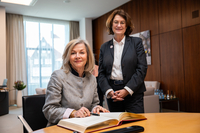 Die Botschafterin sitzt am Besprechungstisch im Präsidentinnenzimmer und trägt sich in das vor ihr liegende Gästebuch ein. Präsidentin Grotheer steht neben ihr. Beide gucken freundlich in die Kamera.