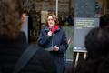 Antje Grotheer steht zwischen zwei Staffeleien und spricht zu den Anwesenden. Sie hält ein Mikrofon in der Hand.