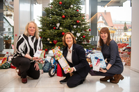 Susanne Eilers, Antje Grotheer und Sylvia Schikker knieben nebeneinander vor derm Wunschbaum. Alle halten ein verpacktes Geschenk in ihren Händen.