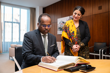 Der Botschafter sitzt am Konferenztisch im Präsidentinnenzimmer und trägt sich in das vor ihm liegende Gästebuch ein. Die Vizepräsidentin steht neben ihm.