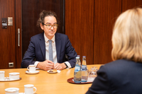 Kieran Drake sitzt am Besprechungstisch im Präsidentenzimmer. Er trägt einen dunklen Anzug. Vor ihm auf dem Tisch stehen eine Kaffeetasse und ein kleiner Teller mit Gebäck.