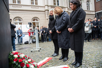 Christine Schnittker, Antje Grotheer und Sahhanim Görgü-Philipp stehen mit gefalteten Händen andächtig vor dem Mahnmal. Vor ihnen auf dem Boden liegt ein Kranz mit Schleife sowie weißen und roten Rosen. Im Hintergrund stehen Zuschauer.