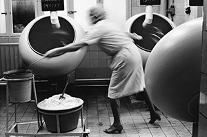 Schwarz-weiß Fotografie: Ein gefliester Raum, in dem mehrere rotierende kugelförmige Maschinen stehen. Eine Frau bewegt sich zwischen zwei der Maschinen. In ihrer linken Hand trägt sie einen großen Löffel, der ein dunkles Pulver enthält. Ihre Darstellung